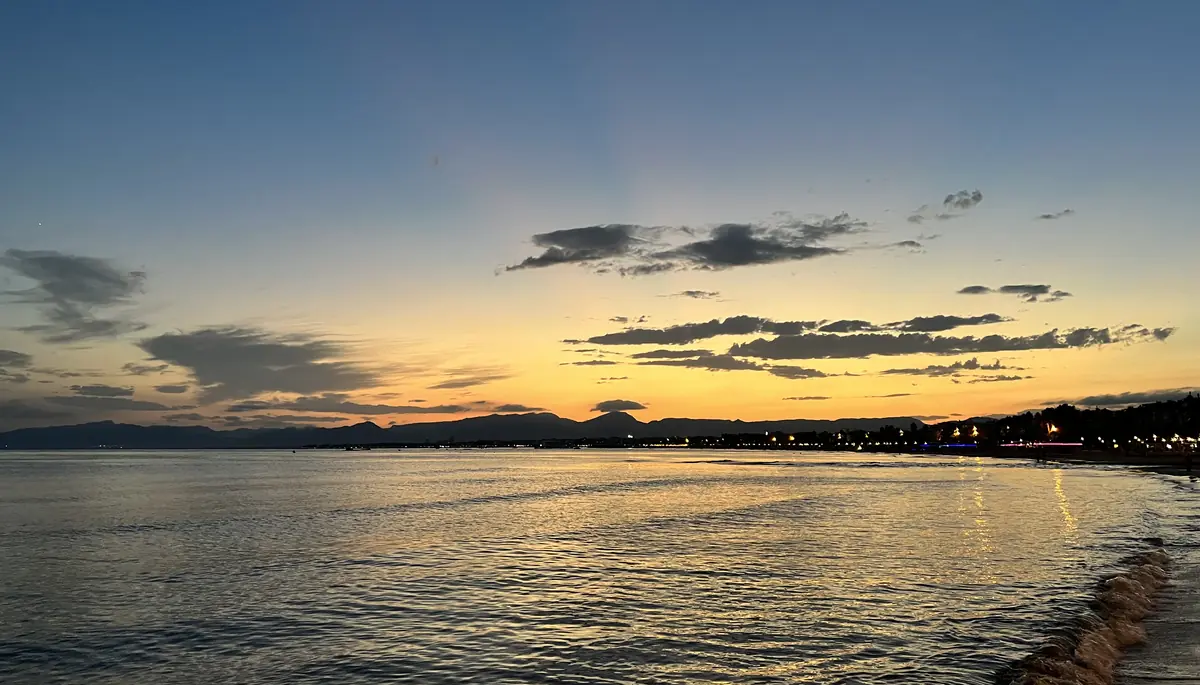 Ein ruhiger Meeresspiegel spiegelt einen gelb-orangen Sonnenuntergangshimmel wider, mit der beleuchteten Küstenlinie von Salou am rechten Rand
