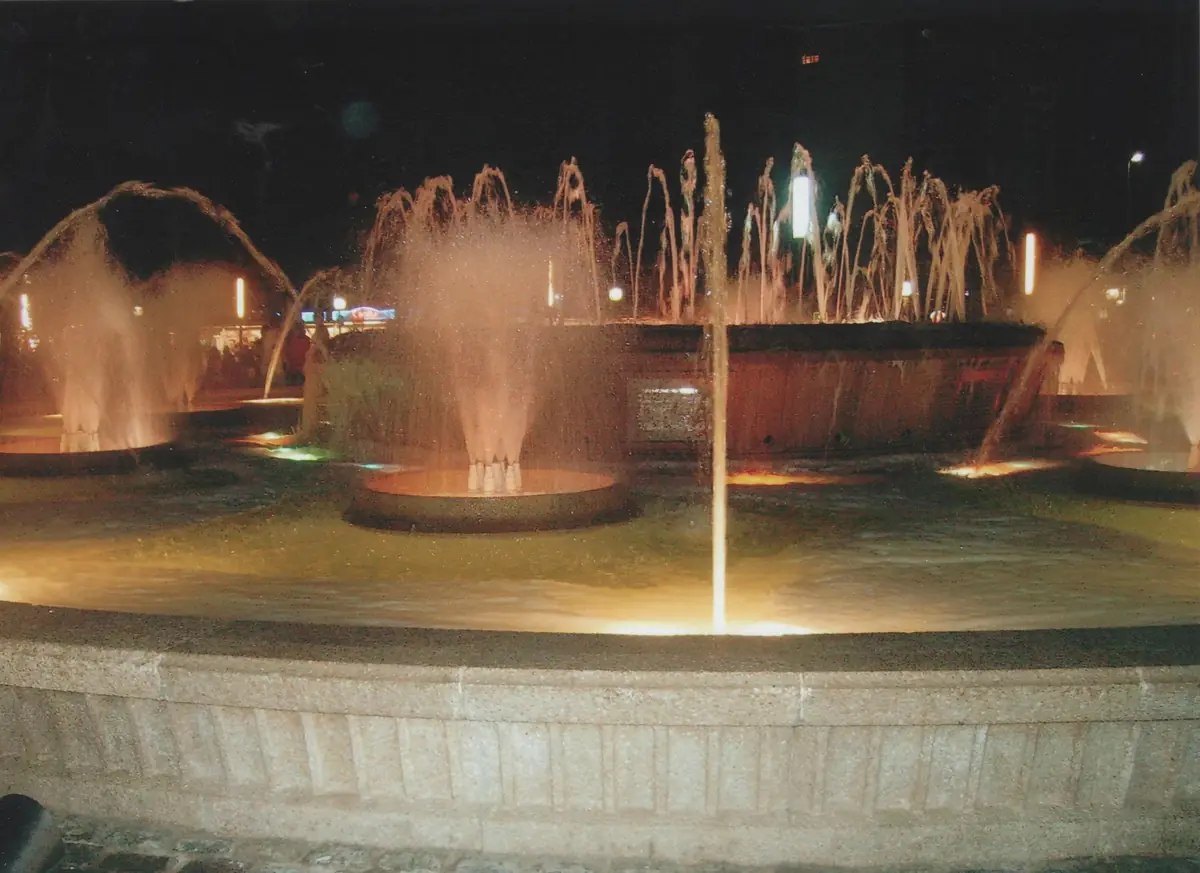Plusieurs jets d'eau s'élèvent d'une fontaine ronde éclairée d'orange contre un ciel nocturne sombre, avec un rebord de pierre bas au premier plan