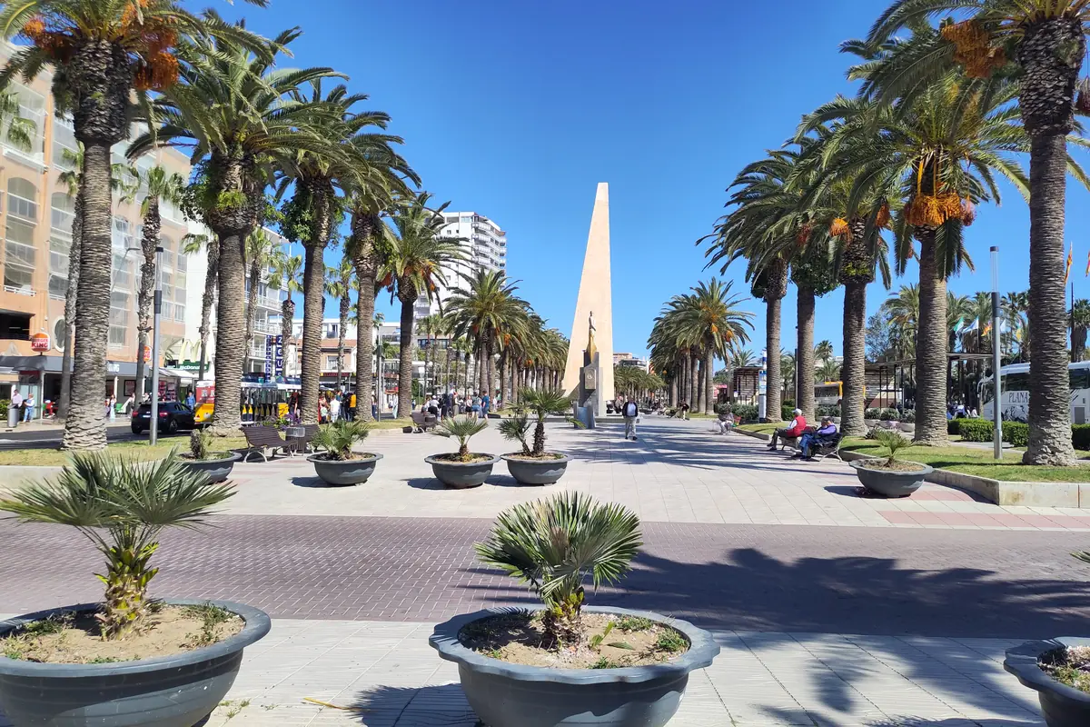 Une large promenade pavée bordée de grands palmiers et de jardinières conduit vers un obélisque haut et élancé sous un ciel bleu