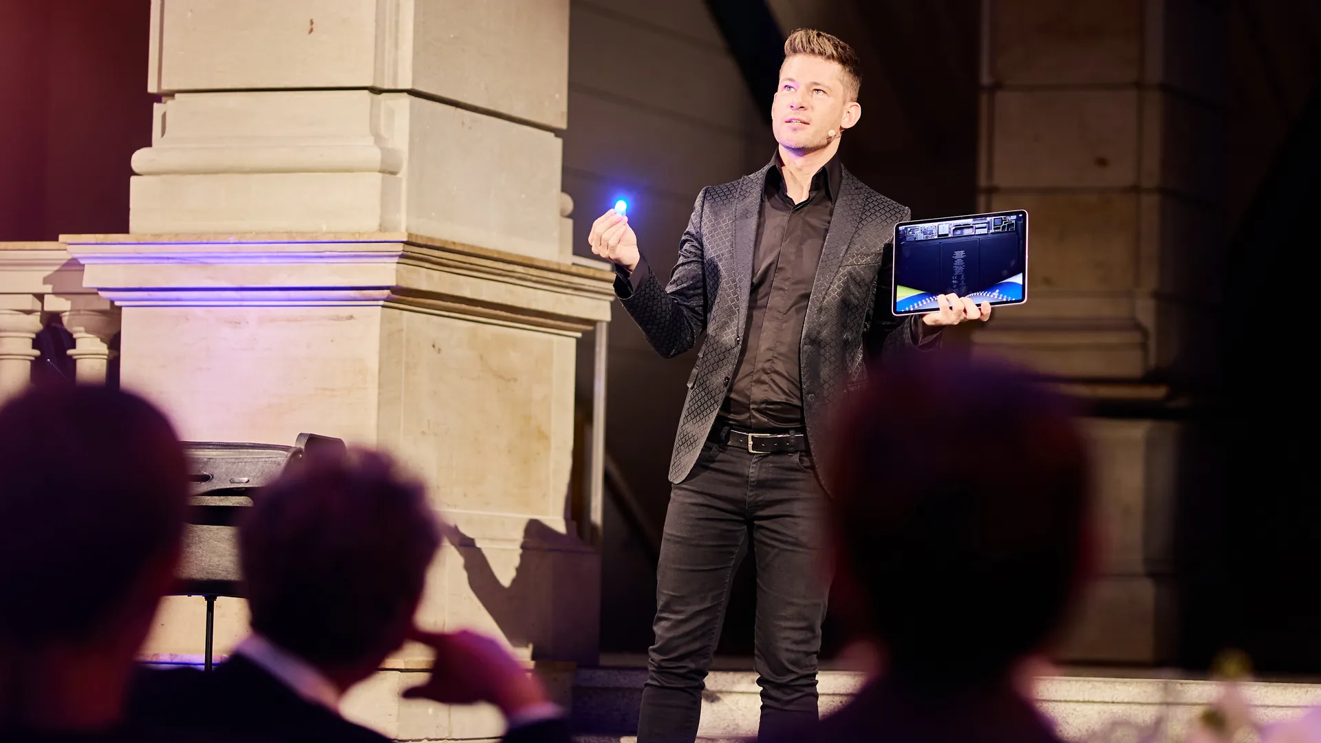 Un mago de escenario con una chaqueta oscura estampada sostiene una luz azul brillante en una mano y una tableta en la otra, con las cabezas del público en primer plano