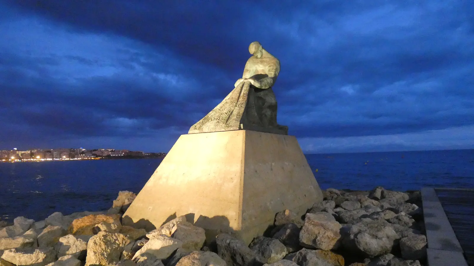 El Monumento a los Pescadores de Salou, una figura de bronce sobre una pirámide de piedra en las rocas del espigón, con las luces de la ciudad reflejadas en el agua bajo un cielo de un azul profundo al caer la tarde
