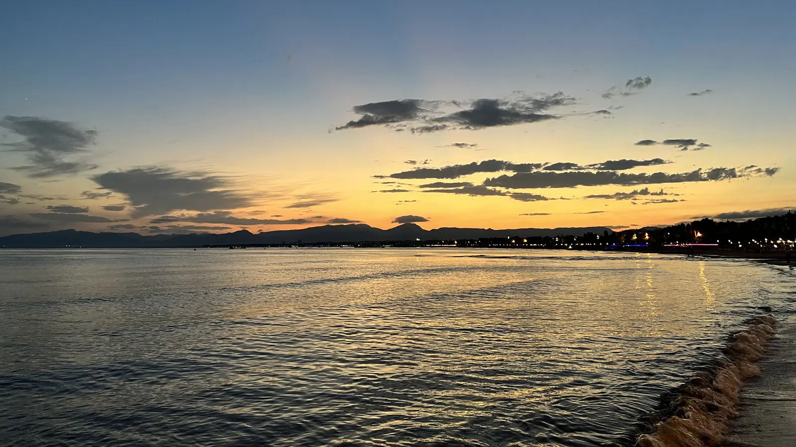 El crepúsculo sobre el mar en Salou, con un cielo naranja y amarillo sobre las aguas en calma y la costa iluminada a la derecha