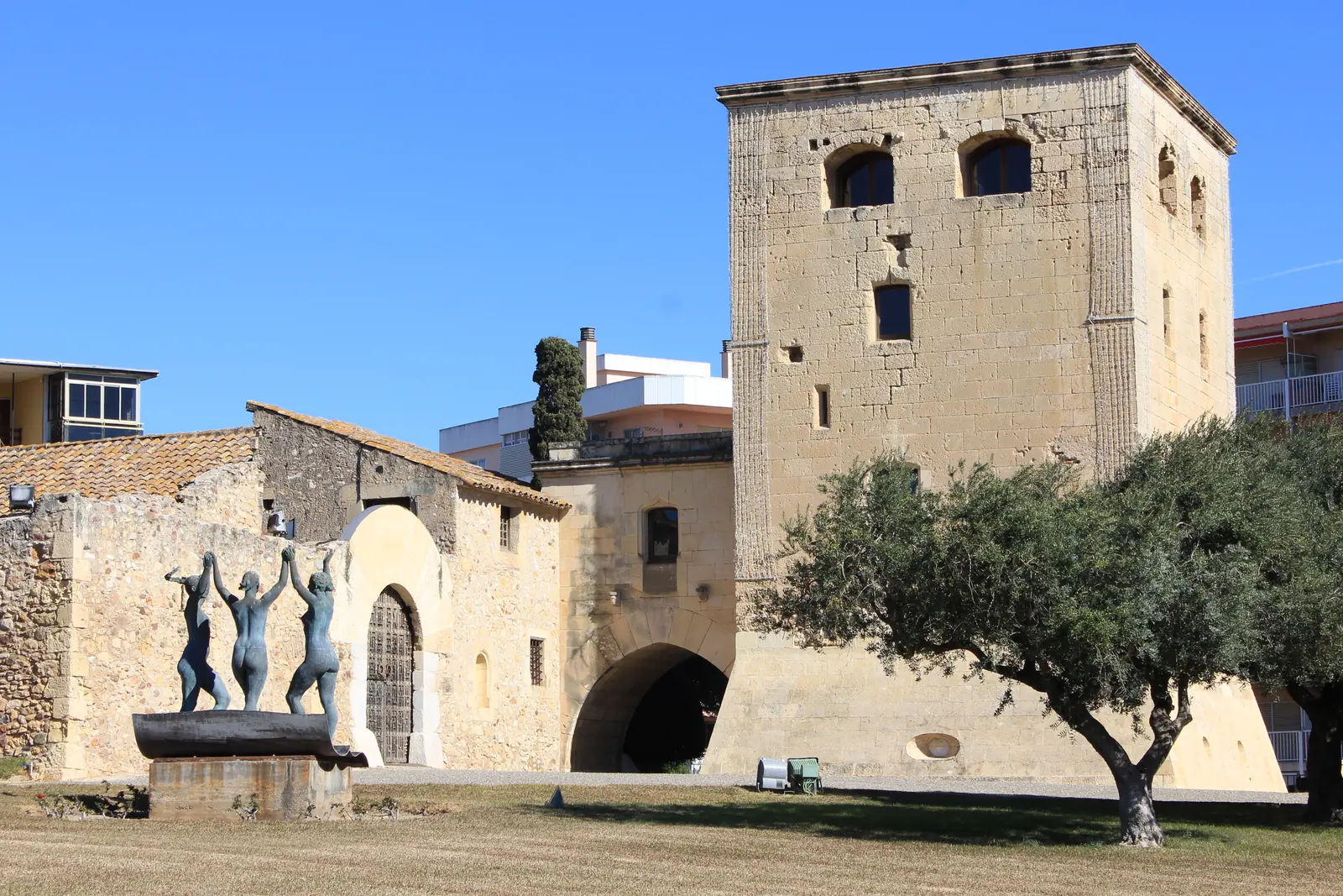 La Torre Vella de Salou, una torre cuadrada de piedra junto a un edificio de piedra, con una escultura de tres figuras frente a ella bajo un cielo azul